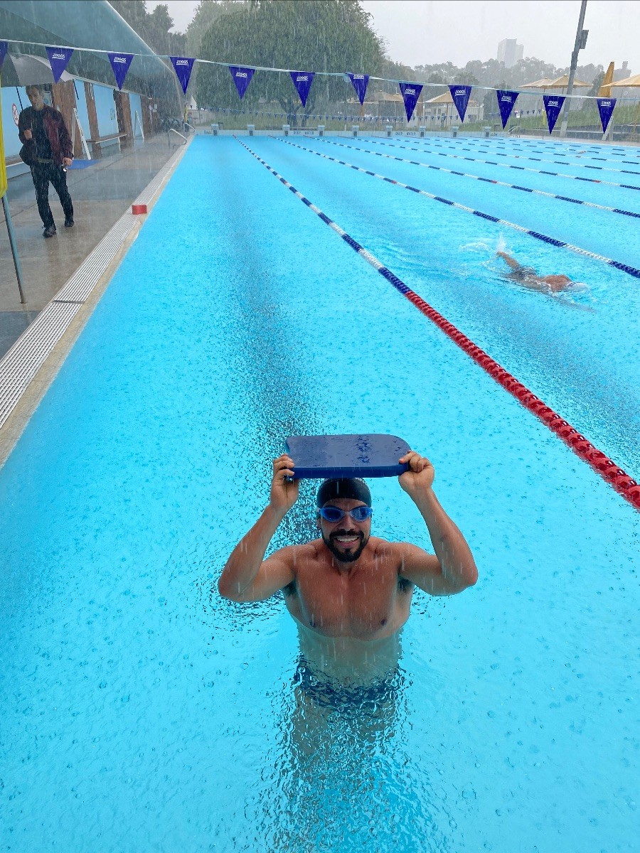 Man standing in pool holding kick board above his head to block out the heavy rain.